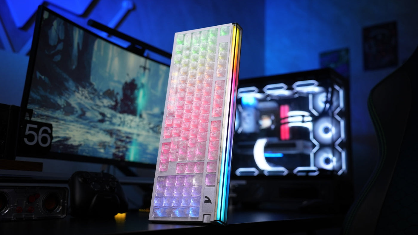 A white translucent RGB keyboard standing on a desk beside a gaming monitor and illuminated PC case